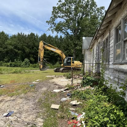 Excavator beside old wooden house in overgrown yard.