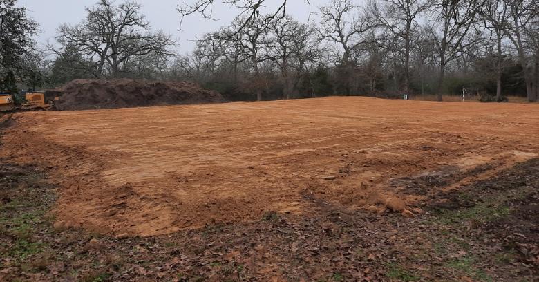 Cleared and leveled dirt construction site surrounded by trees with equipment in the background.